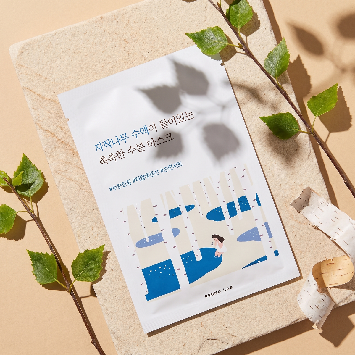 Round Lab Birch Juice Maske liegt im Sonnenlicht auf hellem Sandstein, daneben Birkenzweige und Birkenrinde