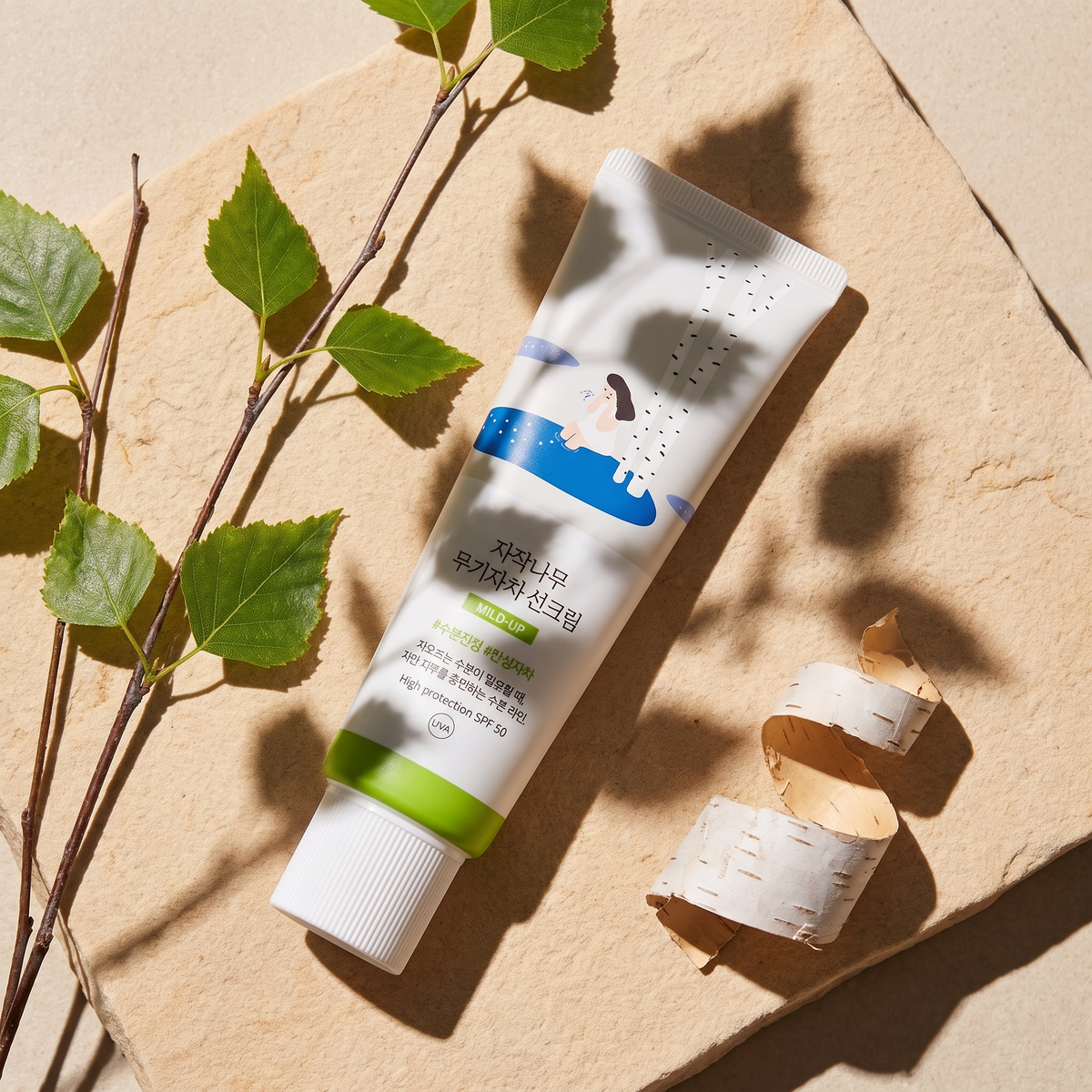 Round Lab Birch Juice Mild Up Sonnencreme liegt im Sonnenlicht auf hellem Sandstein, daneben Birkenzweige und Birkenrinde
