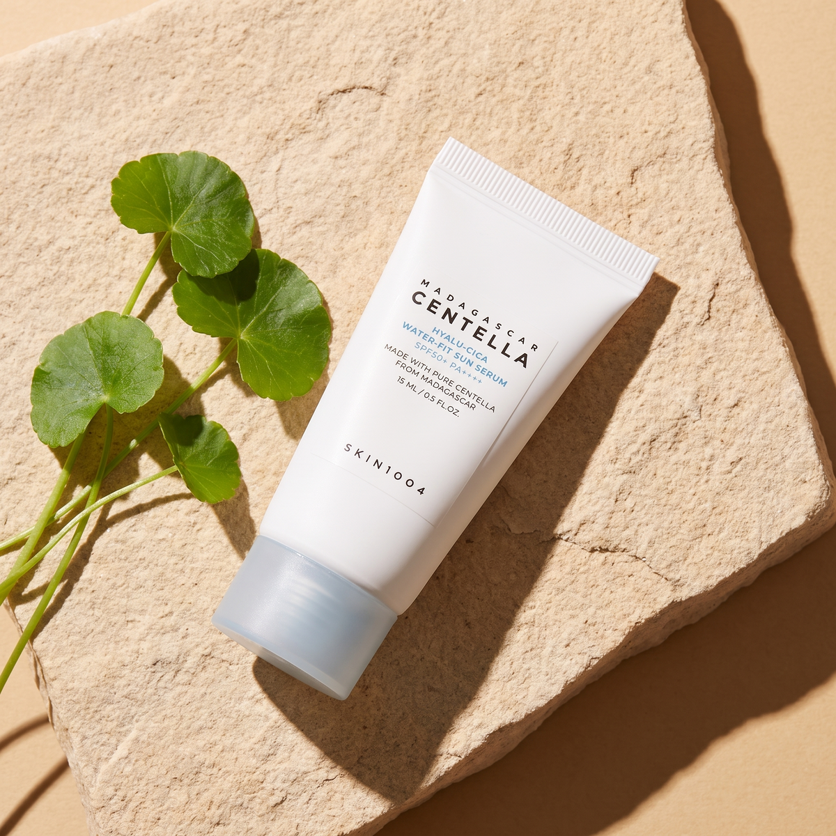 SKIN1004 Hyalu-Cica Water-Fit Sun Serum liegt im Sonnenlicht auf hellem Sandstein, daneben frische Centella-Blätter