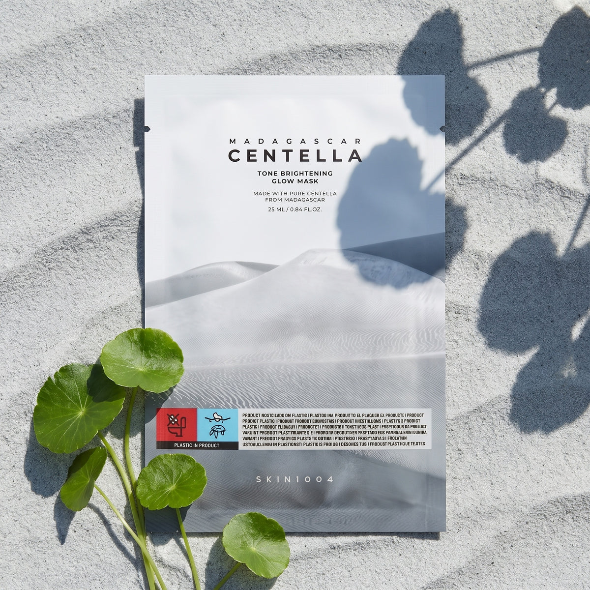 SKIN1004 Tone Brightening Glow Mask liegt auf feinem grauem Sand mit Windrippeln, daneben Centella-Blätter